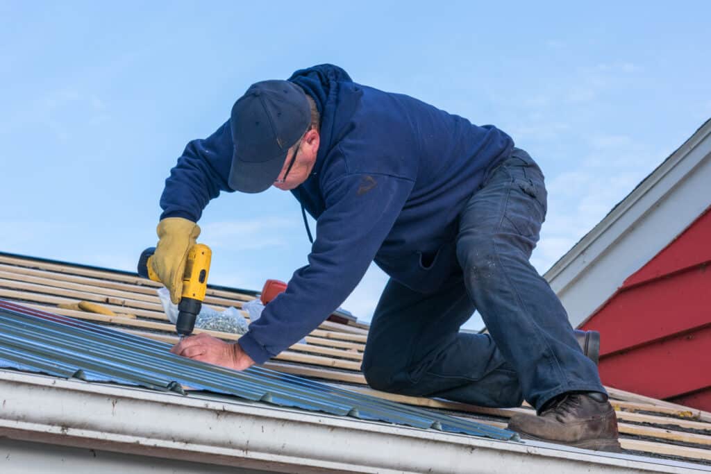 Couvreur installe une toiture métallique ondulée Couvreur avec perceuse jaune fixant des panneaux de toit en métal bleu sur une maison, en plein jour.