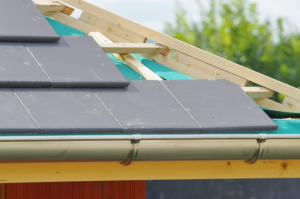 Toiture en construction avec tuiles grises posées, membrane d'étanchéité verte, charpente bois et gouttière métallique installée.
