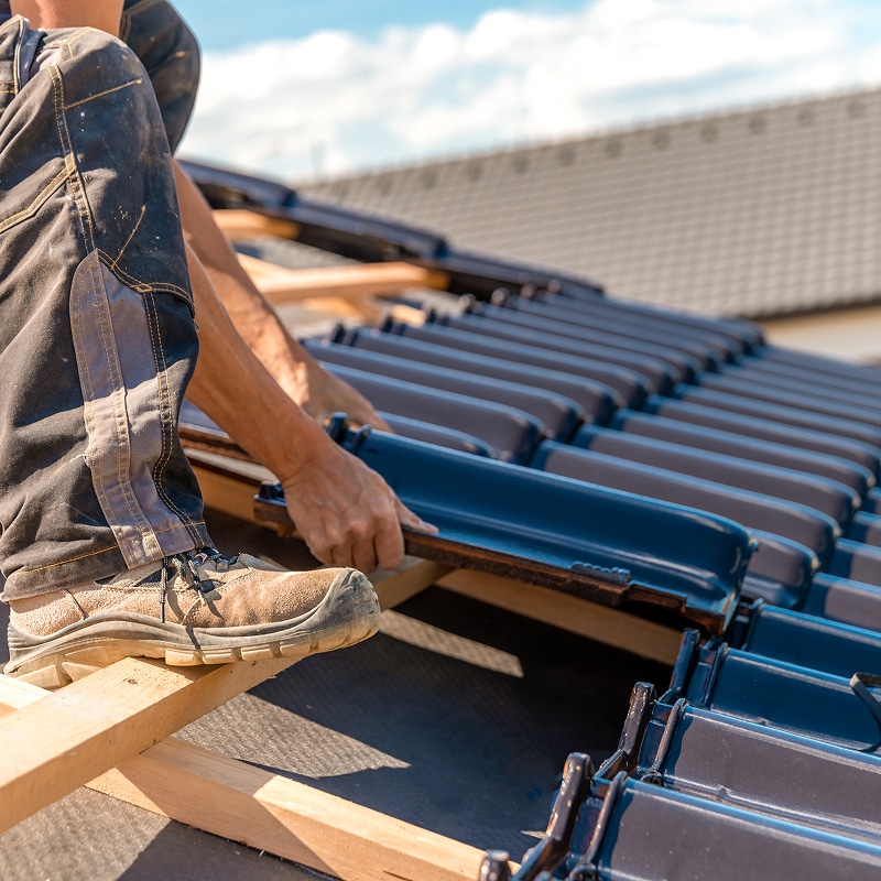 Couvreur installant des tuiles de toit noires sur une toiture en construction, avec des chaussures de sécurité et un ciel bleu.