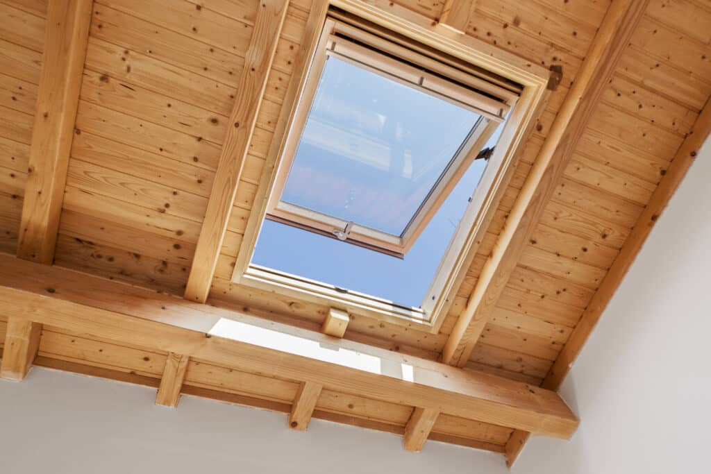 Plafond en bois clair avec poutres apparentes et grande fenêtre de toit ouverte sur un ciel bleu vif, inondant le grenier de lumière naturelle.