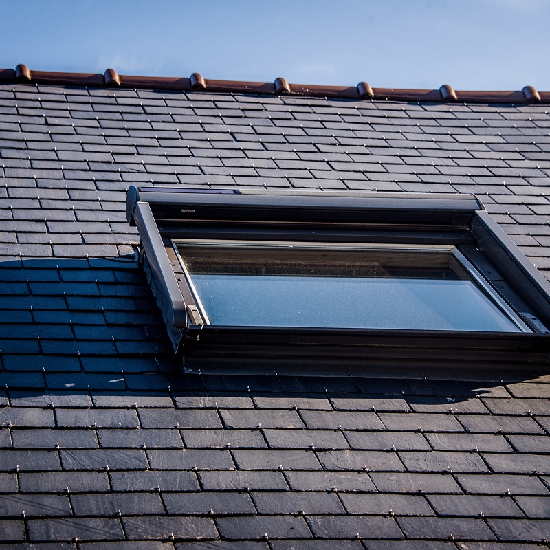 Fenêtre de toit Velux sur un toit en ardoise foncée sous un ciel bleu. Cadre noir et tuiles de faîte marron.
