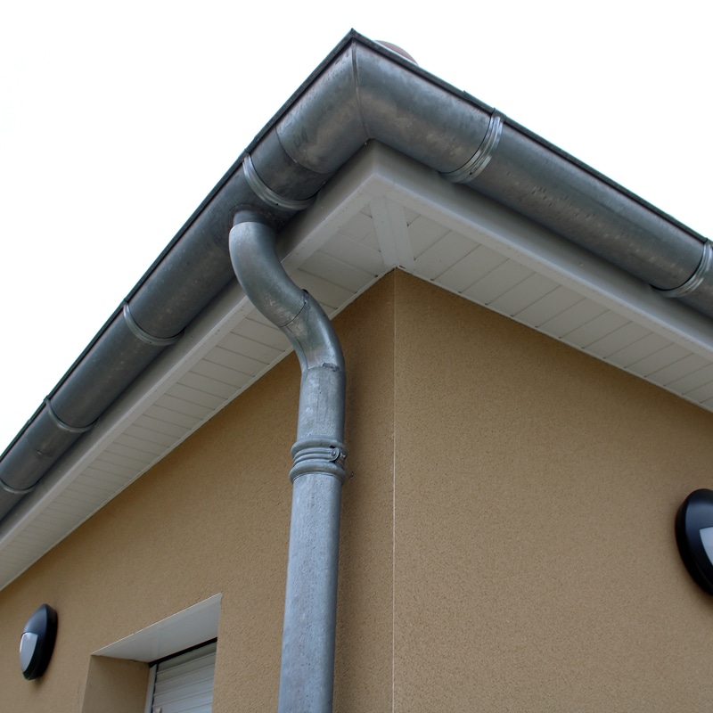 Angle de bâtiment moderne avec gouttière et descente pluviale en métal gris. Façade beige texturée et soffite blanc.