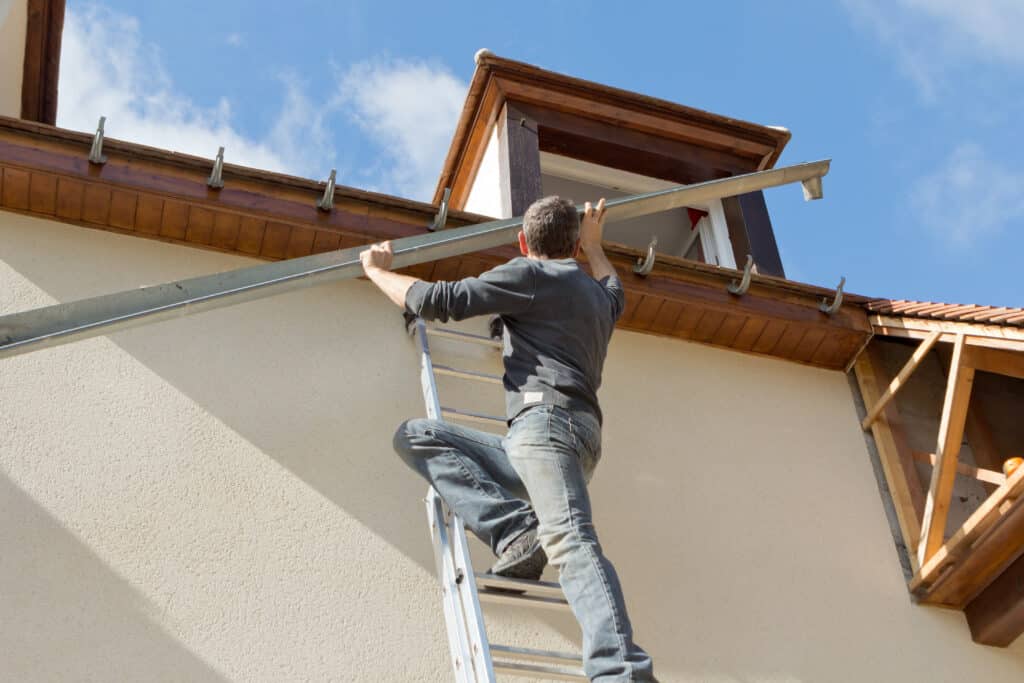 Homme sur échelle posant une gouttière métallique sur la façade d'une maison en bois et crépi, sous un ciel bleu.
