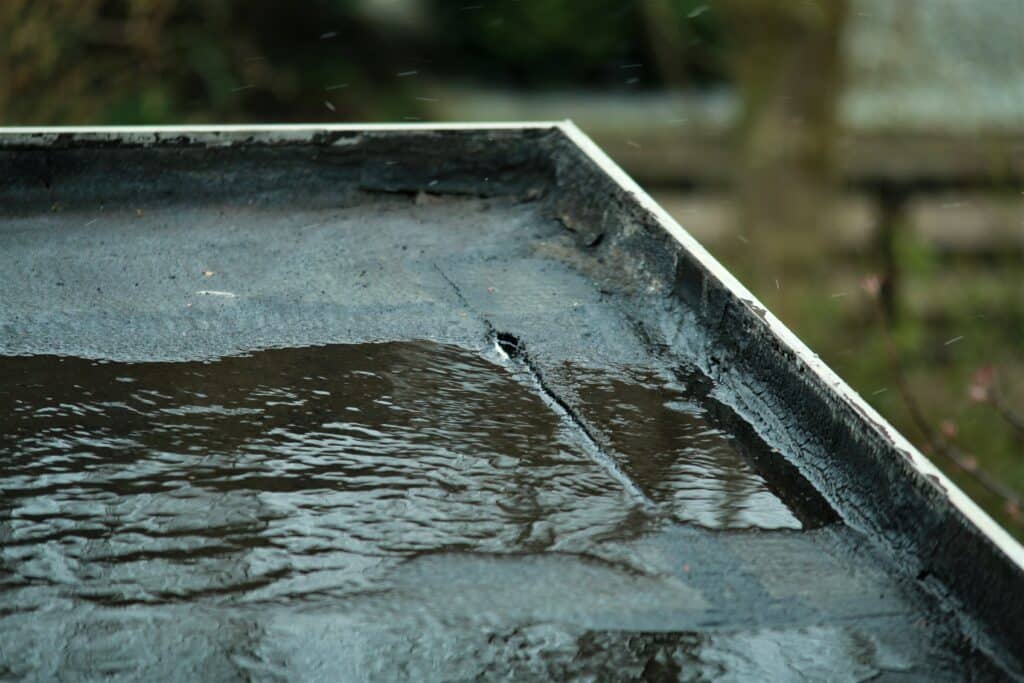 Pluie sur un toit plat : eau stagnante et fissures visibles Surface sombre et fissurée d'un toit plat, recouverte d'eau stagnante et d'ondulations, sous une pluie fine. Arrière-plan flou vert.