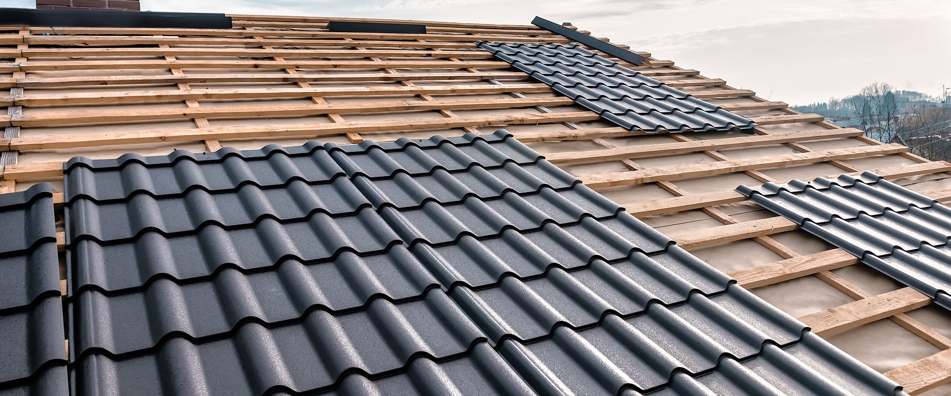Toit en construction avec charpente en bois et pose de tuiles métalliques gris foncé, sous un ciel nuageux.