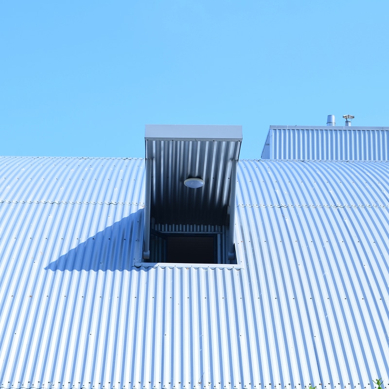Toit incurvé en tôle ondulée argentée sous ciel bleu. Un puits de lumière ouvert avec luminaire et son ombre marquée.