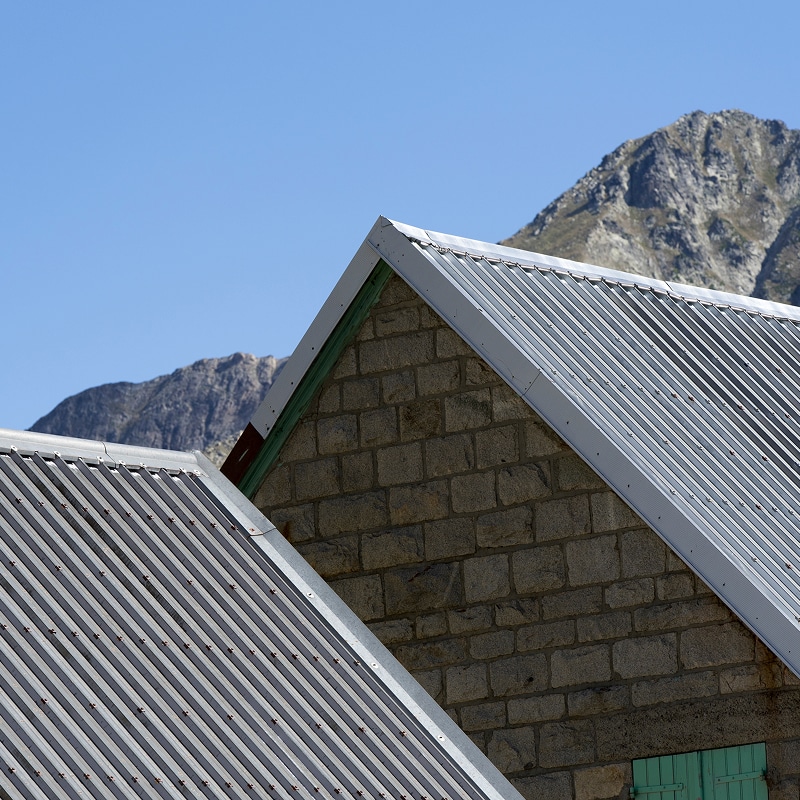 Deux toits en tôle ondulée sur des bâtiments en pierre de montagne, ciel bleu et sommets rocheux en arrière-plan.