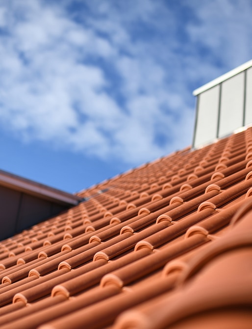 Toit en tuiles de terre cuite orange, nettes et alignées, sous un ciel bleu parsemé de nuages blancs. Architecture moderne.